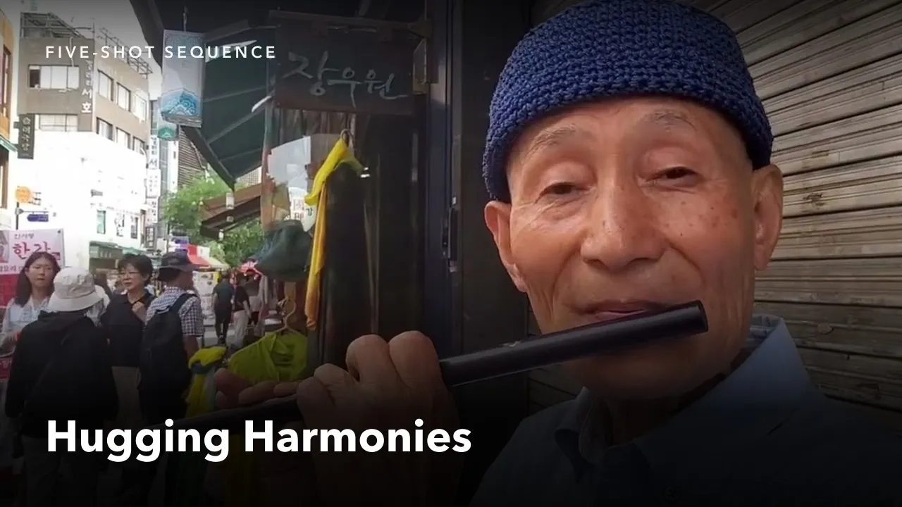 Copertina del video: un uomo anziano con un berretto di lana blu e una camicia azzurra suona un flauto traverso nero, ripreso da vicino in una strada di Insadong, fra le bancarelle e le insegne in coreano. Sopra, l'etichetta «FIVE-SHOT SEQUENCE» e il titolo «Hugging Harmonies».