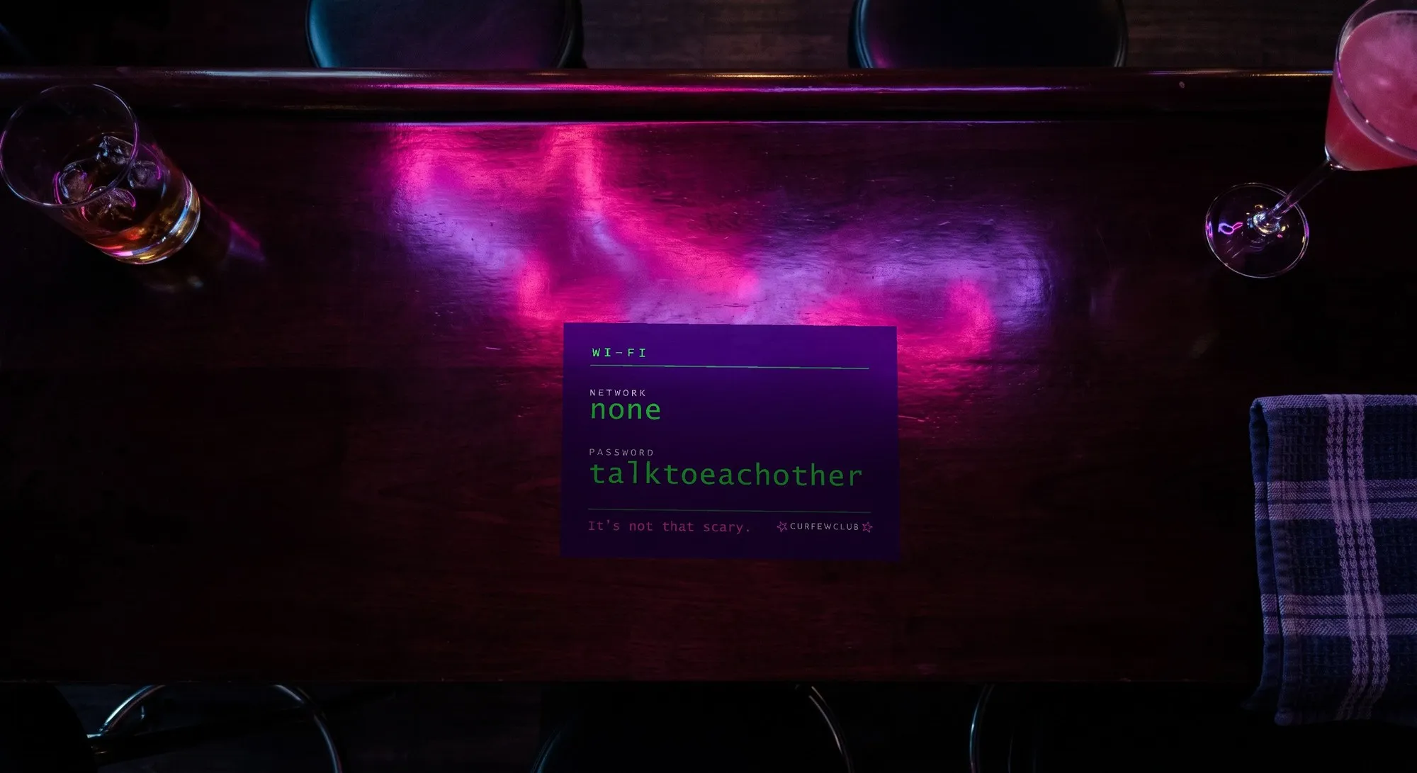 Il bancone scuro di un bar visto dall'alto, con riflessi magenta e viola. Al centro, appoggiata, una targhetta orizzontale che dice «NETWORK: none» e «PASSWORD: talktoeachother» in verde su fondo scuro.