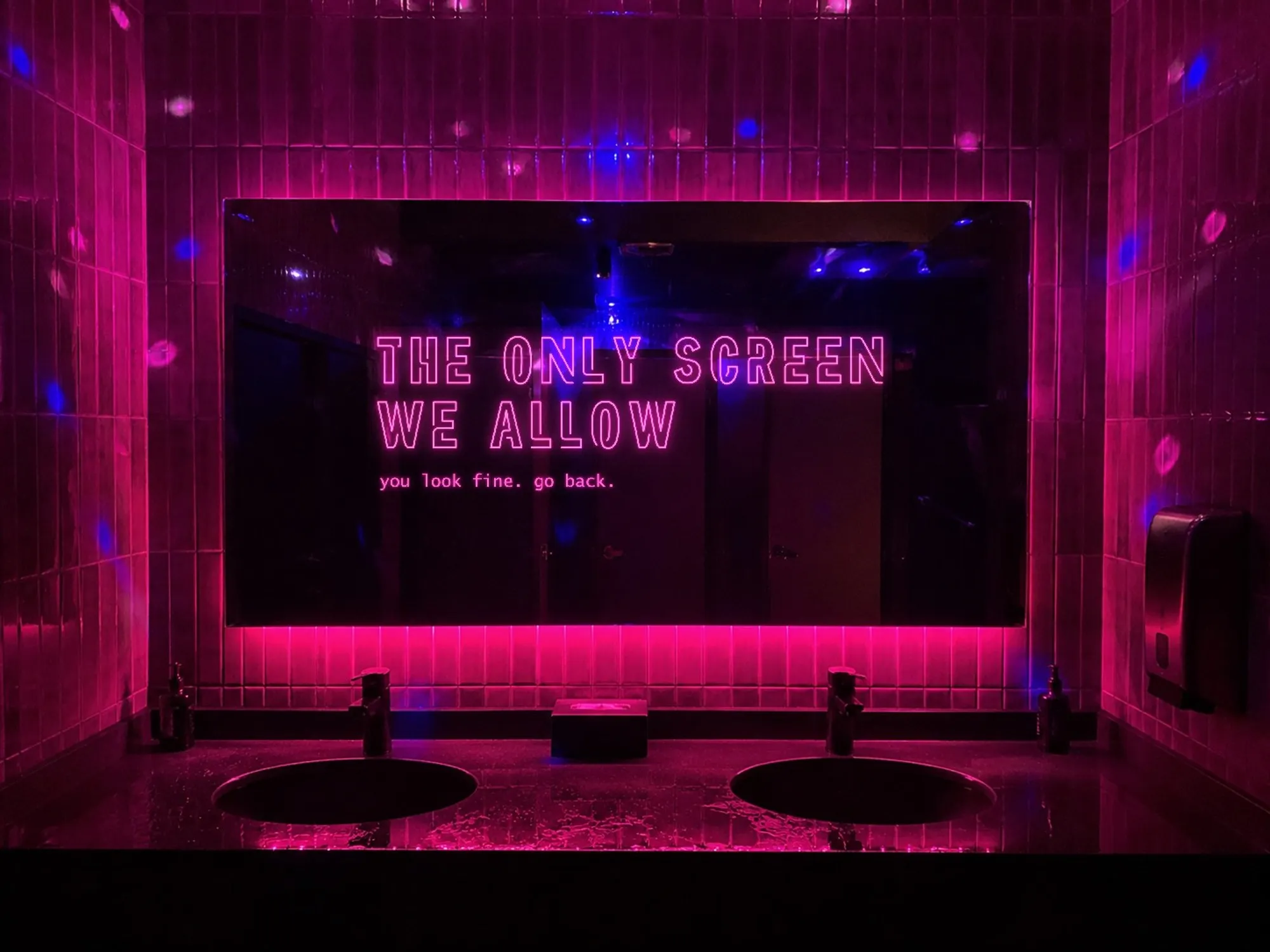 Lo specchio sopra i lavabi di un bagno di locale, con le piastrelle magenta intorno e una striscia di LED rosa sotto. Sul vetro, accesa, un'insegna al neon rosa su due righe — «THE ONLY SCREEN WE ALLOW» — e sotto, più piccolo, «you look fine. go back.»